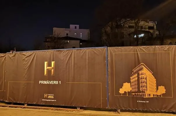 O barieră de construcție maro închis cu logo-ul H și textul PRIMAVERI 1, alături de o ilustrație a unei clădiri.