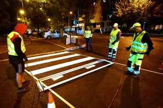 Trei lucrători în veste reflectorizante aplică marcaje rutiere albe pe un carosabil asfaltat noaptea.