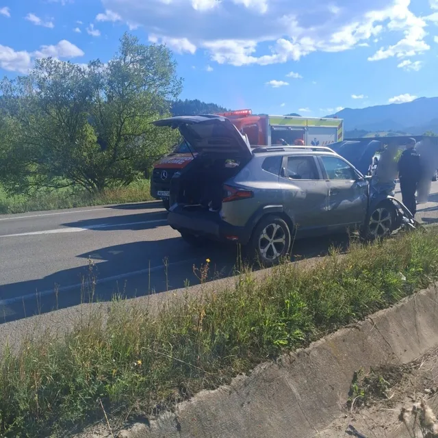 Un autoturism gri cu portbagajul deschis este oprit pe marginea unui drum asfaltat lângă un vehicul de serviciu colorat sub un cer înnorat.