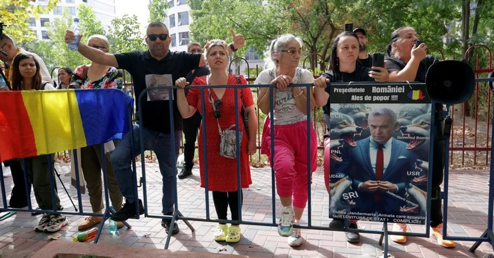 Un grup de persoane stă în spatele unui gard metalic, în timp ce un individ ridică un poster cu un bărbat în costum și cravată roșie.