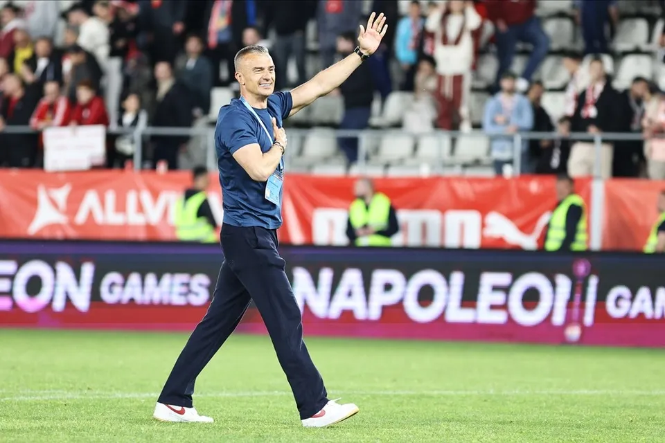 Un bărbat cu tricou albastru și pantaloni închiși merge pe un teren de fotbal, ridicând un braț spre tribunele cu spectatori din fundal.