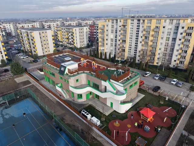 O clădire modernă cu fațadă verde și albă este situată lângă un teren de sport și blocuri de locuințe înalte sub un cer înnorat.