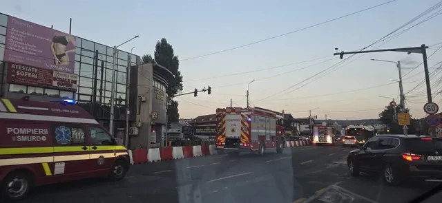 Vehicule de serviciu de culoare roșu și alb, cu lumini de alertă aprinse, sunt parcate pe un drum asfaltat sub un cer senin.