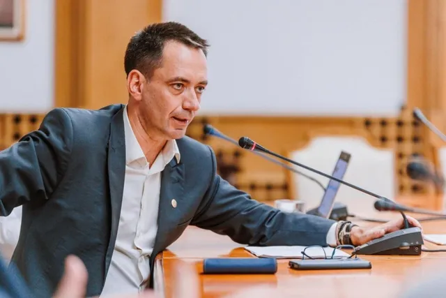 Un bărbat cu sacou și cămașă albă stă la o masă de conferință lângă microfoane și un telefon fix.