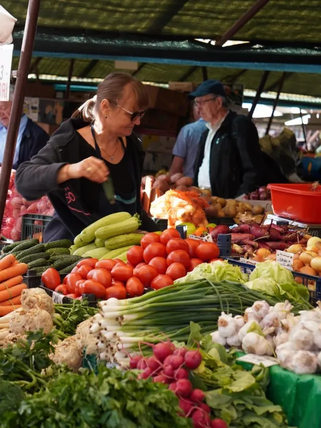O femeie cu ochelari stă în spatele unui stand de legume și fructe, lângă un bărbat cu șapcă și ochelari.