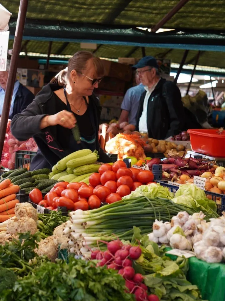 O femeie cu ochelari stă în spatele unui stand de legume și fructe, lângă un bărbat cu șapcă și ochelari.