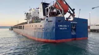 O navă mare, de culoare albastră cu partea inferioară roșie, pe care scrie CHELSEA-3 VALLETTA, navighează pe apă lângă un mal.