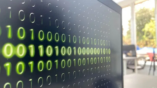 Linii de cifre binare verzi sunt afișate pe un ecran de computer întunecat, în timp ce fundalul este neclar și luminos.