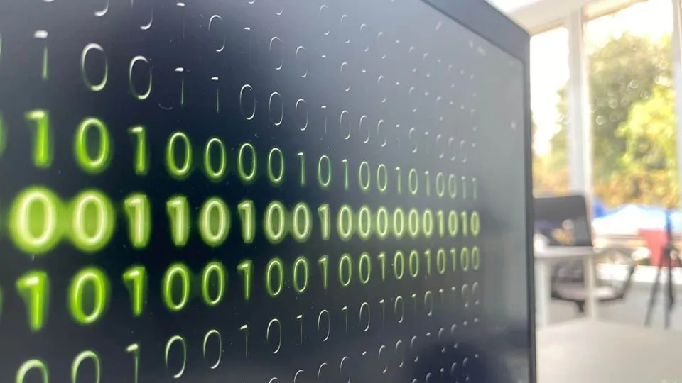 Linii de cifre binare verzi sunt afișate pe un ecran de computer întunecat, în timp ce fundalul este neclar și luminos.