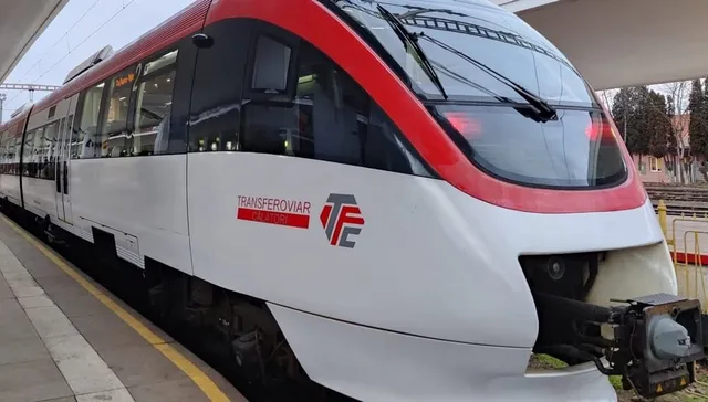 Un tren alb și roșu, cu logo pe lateral, este staționat lângă o peroană acoperită, cu linii și copaci în fundal.