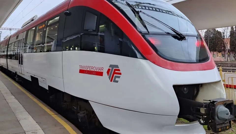 Un tren alb și roșu, cu logo pe lateral, este staționat lângă o peroană acoperită, cu linii și copaci în fundal.