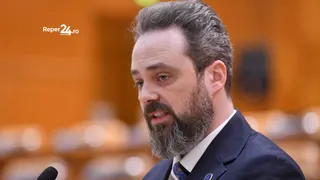 Un bărbat cu barbă și păr scurt, purtând sacou închis și cămașă albă, este surprins într-o poziție de profil.