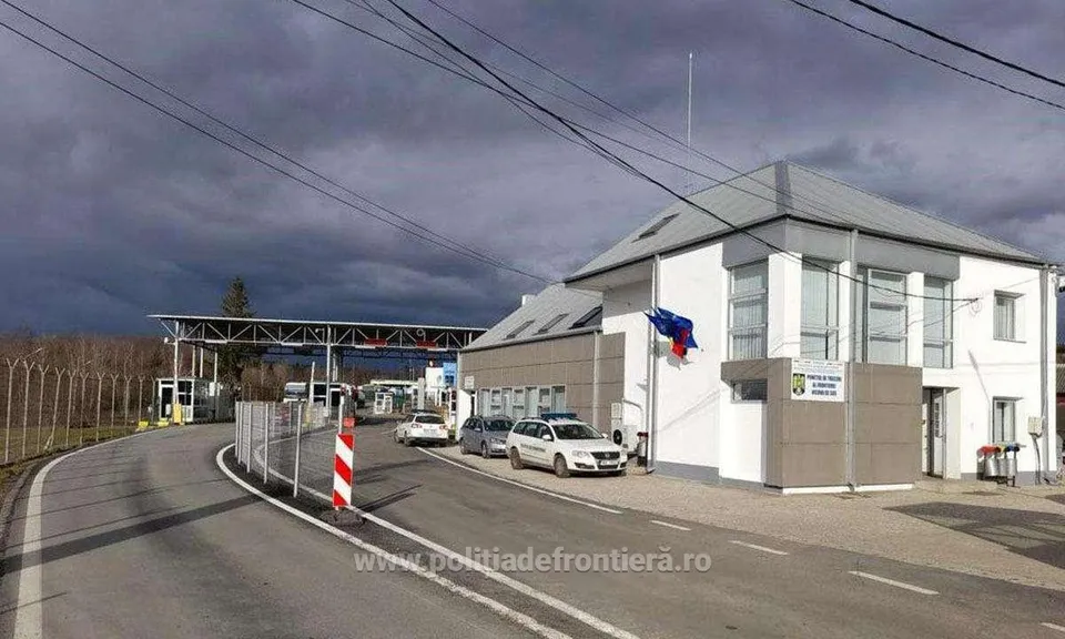 O clădire albă cu acoperiș gri stă lângă un drum asfaltat pe care sunt parcate trei mașini sub un cer înnorat.