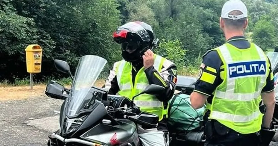 Un agent de poliție în uniformă reflectorizantă stă lângă un motociclist care poartă cască și vestă reflectorizantă lângă o motocicletă neagră pe un drum.