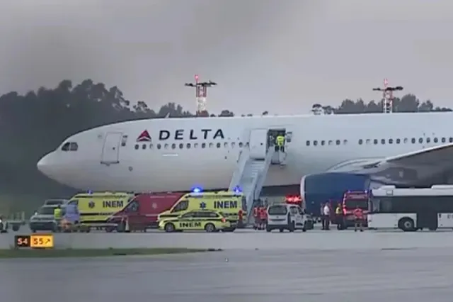 Un avion alb cu inscripția Delta este parcat pe o pistă lângă mai multe vehicule de serviciu de culoare galbenă și roșie.