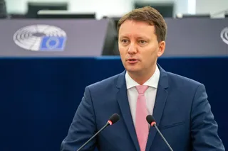 Un bărbat cu părul scurt, purtând sacou albastru, cămașă albă și cravată roz, stă în fața a două microfoane.
