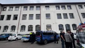 Mașini de poliție și o ambulanță sunt parcate în fața unei clădiri albe cu ferestre multiple, în prezența unor persoane.