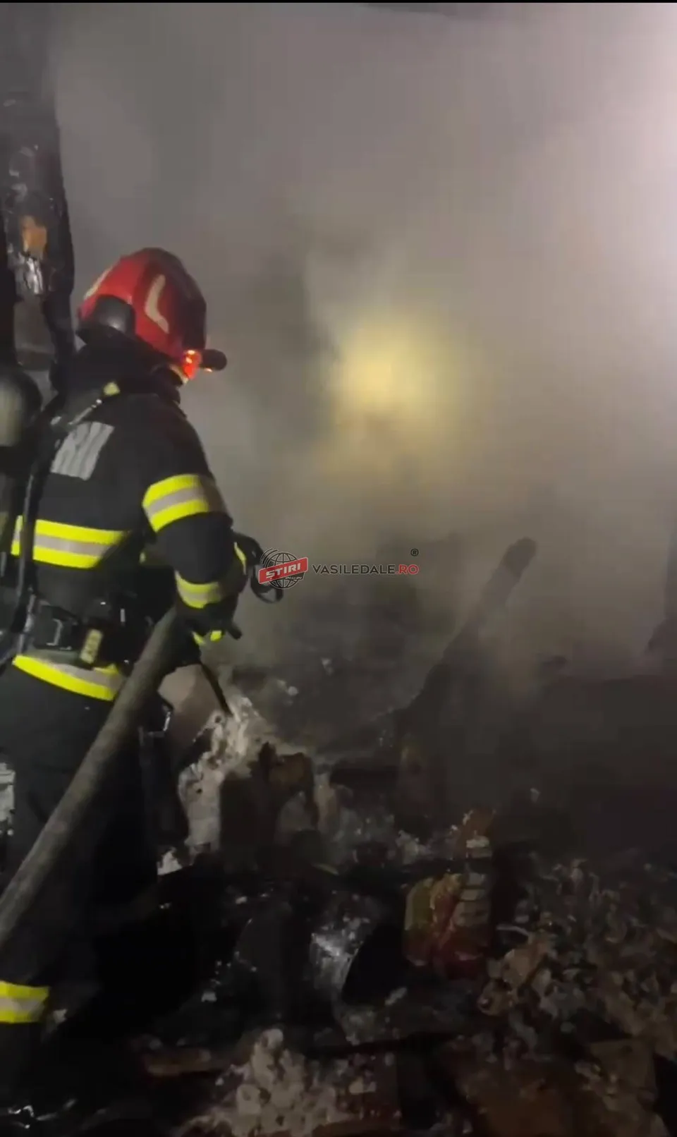 Un pompier cu echipament de protecție și cască este orientat spre o zonă plină de fum și resturi carbonizate.