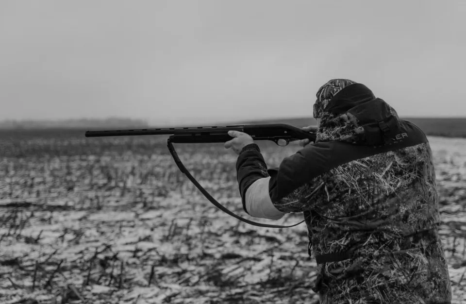 O persoană îmbrăcată în haine de camuflaj țintește o armă de vânătoare spre orizontul unui câmp deschis sub un cer înnorat.