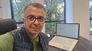 Un bărbat cu ochelari și vestă neagră stă la o masă lângă un laptop deschis într-o cameră cu fereastră mare.