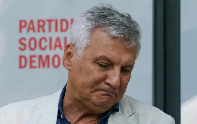 Un bărbat cu părul cărunt și o jachetă deschisă stă cu privirea în jos în fața unui fundal cu text roșu.