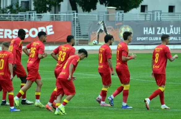 Cei opt jucători de fotbal poartă echipamente roșii și se află pe un teren de gazon verde sub un cer deschis.
