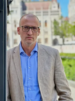 Un bărbat cu ochelari, în costum deschis și cămașă albastră, stă în picioare în fața unei clădiri neclară.