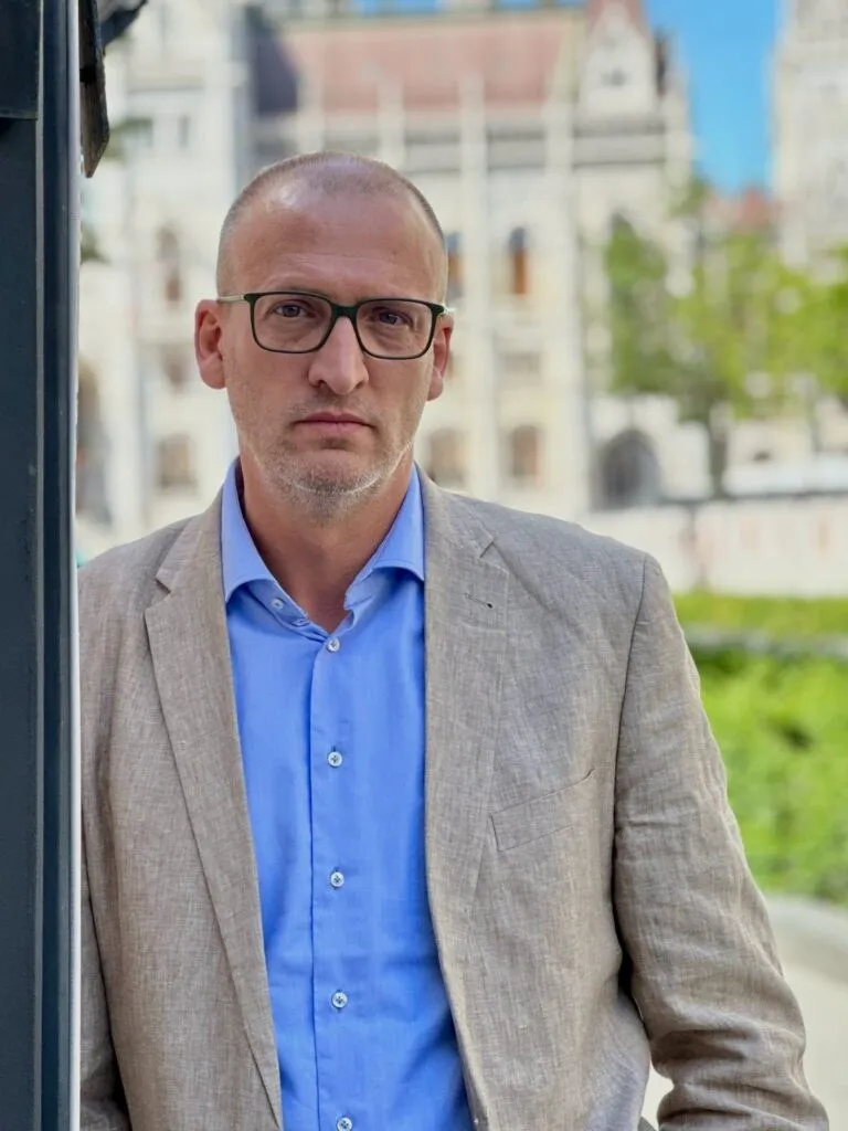 Un bărbat cu ochelari, în costum deschis și cămașă albastră, stă în picioare în fața unei clădiri neclară.
