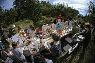 Oamenii stau la mese lungi în aer liber, pictând pe pânze de pe suporturi de lemn înconjurați de copaci și iarbă sub un cer senin.