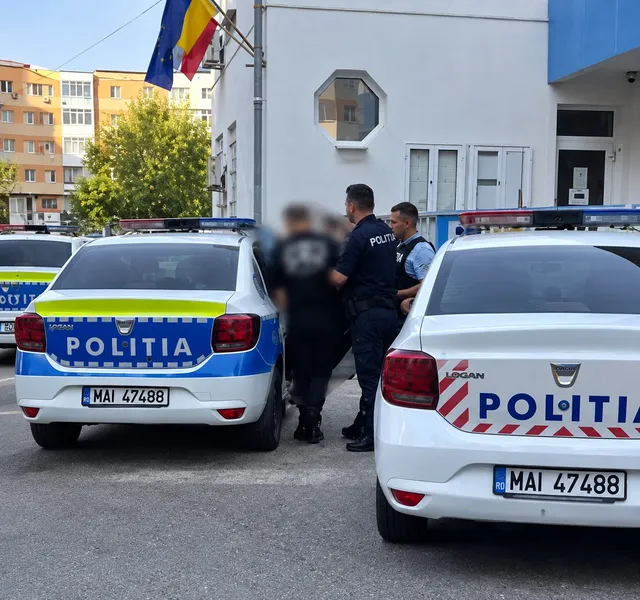 Două mașini de poliție albe cu inscripții albastre și galbene sunt parcate pe un drum, în fața unei clădiri albe, lângă câțiva bărbați în uniformă.