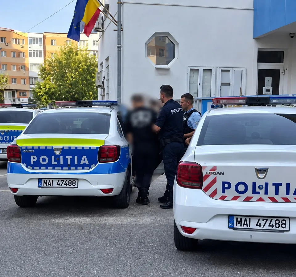 Două mașini de poliție albe cu inscripții albastre și galbene sunt parcate pe un drum, în fața unei clădiri albe, lângă câțiva bărbați în uniformă.