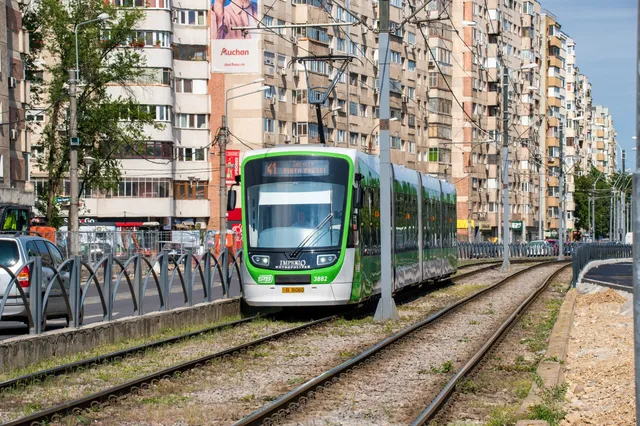 Un tramvai verde circulă pe șine de tramvai într-o zonă urbană cu clădiri înalte de apartamente în fundal.