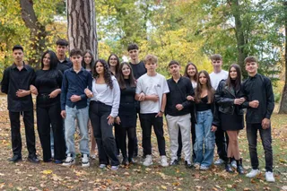 Un grup de tineri stă în rând pe un teren cu iarbă și frunze uscate, în fața unor copaci cu frunze verzi și galbene.