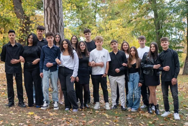 Un grup de tineri stă în rând pe un teren cu iarbă și frunze uscate, în fața unor copaci cu frunze verzi și galbene.