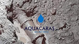 O țeavă metalică este vizibilă într-o șanț de excavare plin cu apă și pietriș, având un logo suprapus în partea dreaptă.