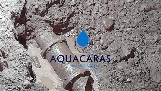 O conductă metalică este vizibilă într-un șanț plin cu apă și pietriș, având un logo suprapus în partea centrală.