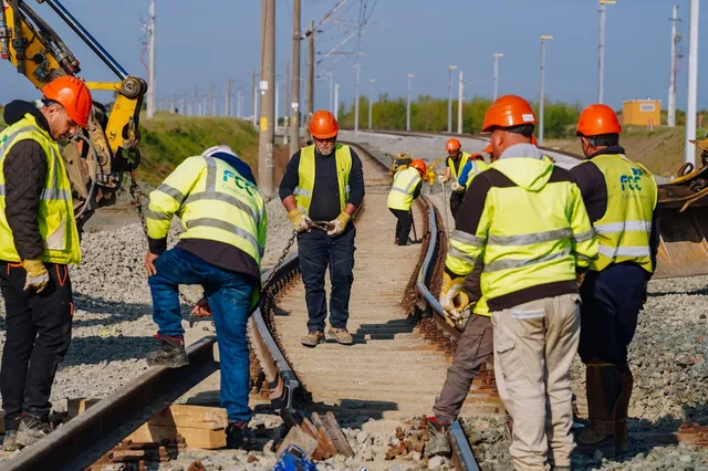 O echipă de lucrători în veste reflectorizante de siguranță lucrează pe o cale ferată lângă un utilaj de excavare sub un cer senin.