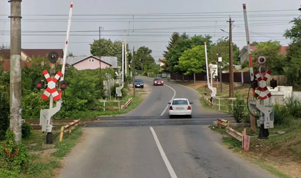 O drum cu o trecere la nivel cu calea ferată, semnale de cale ferată, o mașină albă și altele pe fundal, într-o zonă rurală.
