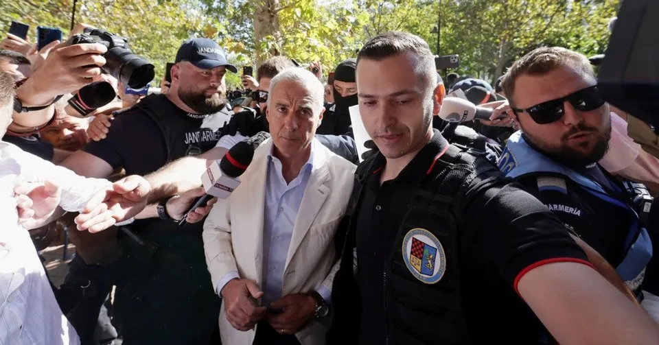 Un bărbat într-un sacou deschis este înconjurat de microfoane și persoane care poartă haine negre și ochelari de soare în exterior.