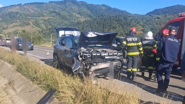 Un vehicul de culoare închisă cu capota deschisă stă pe marginea drumului lângă persoane purtând uniforme de salvatori și poliție.
