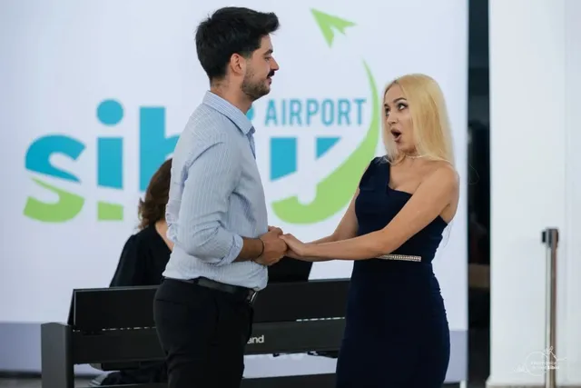 Un bărbat într-o cămașă albastră și o femeie într-o rochie neagră stau față în față în fața unui panou cu un logo colorat.