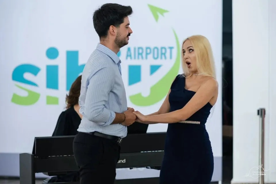 Un bărbat într-o cămașă albastră și o femeie într-o rochie neagră stau față în față în fața unui panou cu un logo colorat.