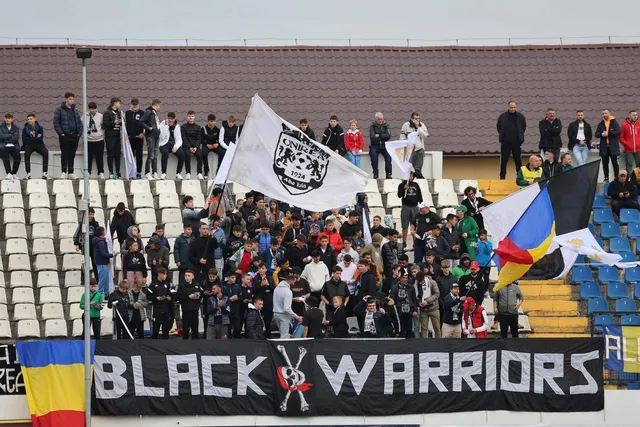 Un grup de oameni stă pe tribunele unui stadion, lângă un banner negru cu textul alb și un steag alb cu un logo central.