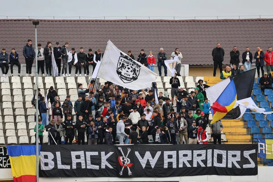 Un grup de oameni stă pe tribunele unui stadion, lângă un banner negru cu textul alb și un steag alb cu un logo central.