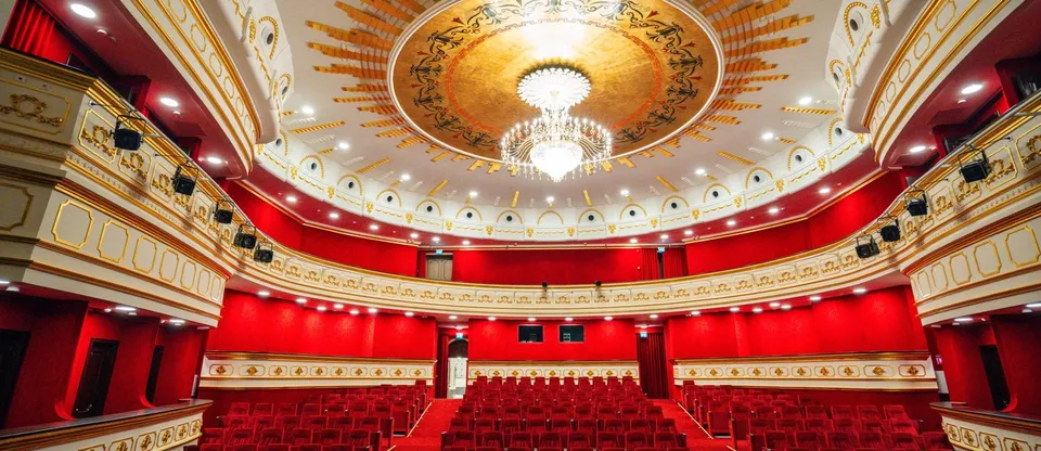 Interiorul unui auditorium cu scaune roșii, pereți roșii și un plafon decorat cu un candelabru central mare și modele aurii.