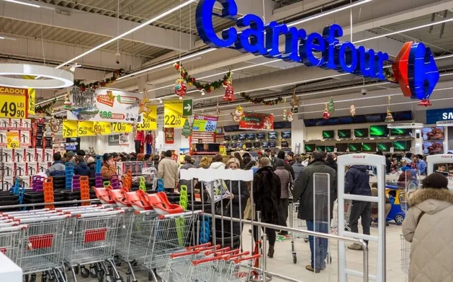 Oamenii stau în rând în interiorul unui magazin, lângă coșuri de cumpărături și un semn albastru suspendat pe tavan.