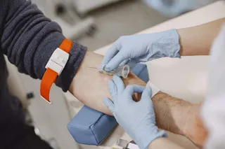 O persoană cu un brățar portocalie își are brațul întins, în timp ce o altă persoană cu mănuși albastre folosește o seringă pentru o procedură medicală.