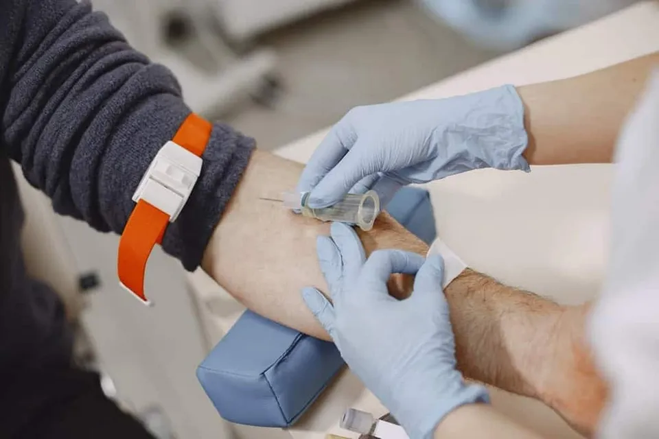 O persoană cu un brățar portocalie își are brațul întins, în timp ce o altă persoană cu mănuși albastre folosește o seringă pentru o procedură medicală.