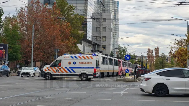 O ambulanță albă cu dungi albastre și galbene stă pe o stradă lângă un tramvai, în fața unor clădiri și copaci.