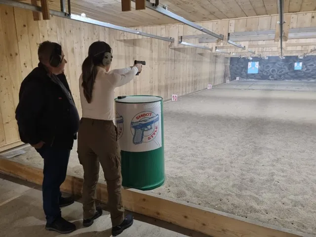 O femeie cu părul lung și o cămașă albă țintește un pistol într-un poligon de tragere, în timp ce un bărbat o privește de lângă ea.